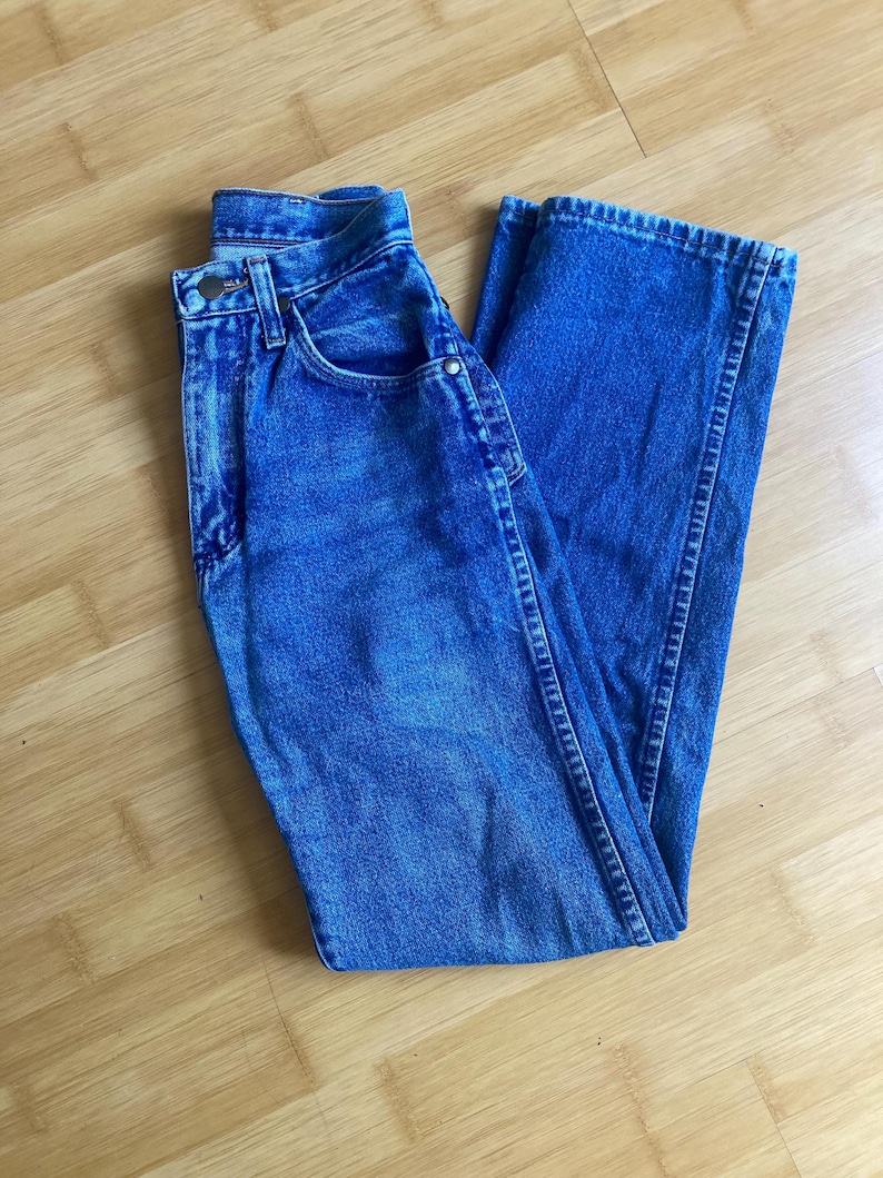 May include: A pair of medium-wash blue denim jeans featuring a high-waisted design, a classic button and zip fly closure, belt loops, and traditional five-pocket styling with metal rivets and visible stitching along the seams.
