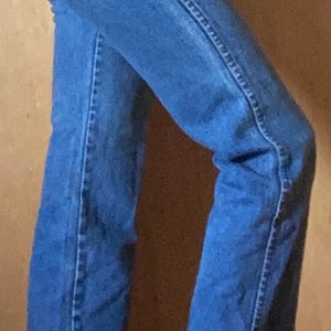 May include: High-waisted blue denim jeans featuring a straight-leg cut, a classic five-pocket design, a metal button closure, and a small rectangular brown leather patch located on the back right pocket area, shown from a side profile view.