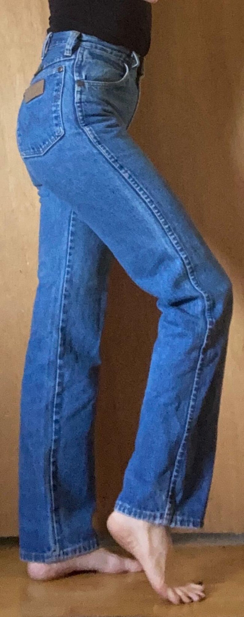 May include: High-waisted blue denim jeans featuring a straight-leg cut, a classic five-pocket design, a metal button closure, and a small rectangular brown leather patch located on the back right pocket area, shown from a side profile view.