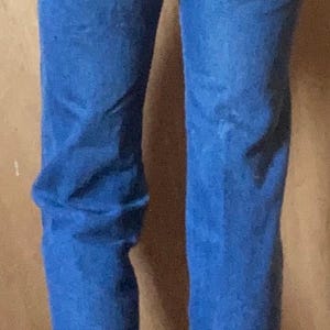 May include: High-waisted blue denim jeans featuring a classic five-pocket design, a leather-like patch on the back right pocket, belt loops, and a button-fly closure. The medium-wash denim fabric shows visible stitching and a straight-leg cut.