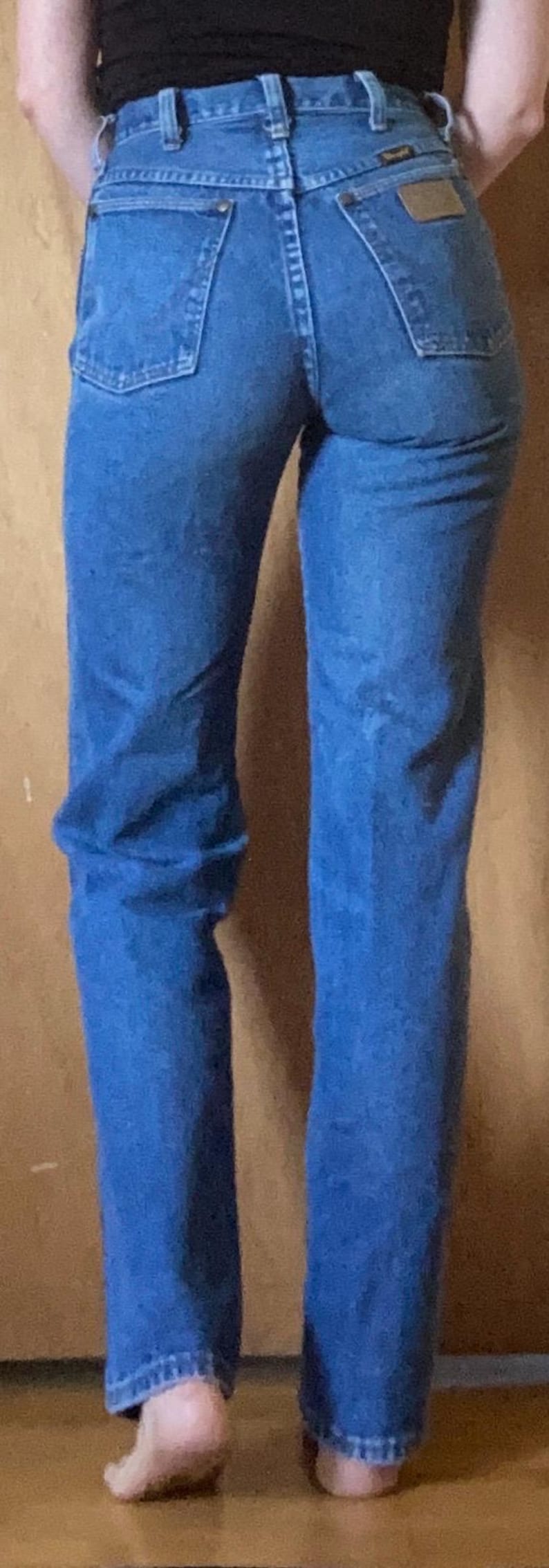 May include: High-waisted blue denim jeans featuring a classic five-pocket design, a leather-like patch on the back right pocket, belt loops, and a button-fly closure. The medium-wash denim fabric shows visible stitching and a straight-leg cut.