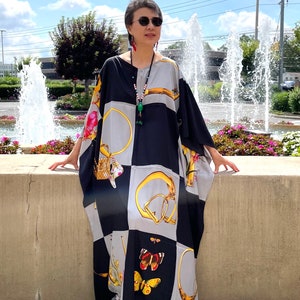 May include: A woman wearing a black and gray patterned maxi dress with gold accents and a butterfly design. She is standing in front of a fountain and wearing sunglasses and a necklace.