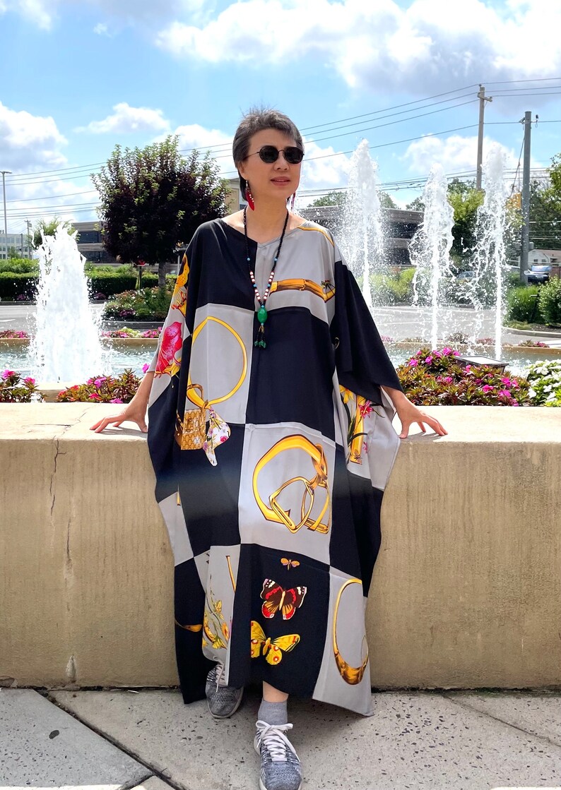 May include: A woman wearing a black and gray patterned maxi dress with gold accents and a butterfly design. She is standing in front of a fountain and wearing sunglasses and a necklace.