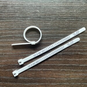 Può includere: Tre misuratori di anelli in plastica bianca con segni neri. I misuratori sono etichettati "SIZE & HALF SIZES" e hanno numeri da 1 a 17. I misuratori sono contrassegnati "PAT. PEND. 9101693".