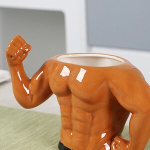 May include: A brown ceramic mug shaped like a muscular torso with raised arms. The mug is sitting on a green table runner.