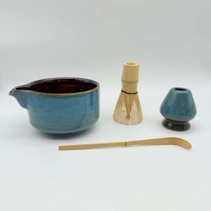 May include: A matcha tea set comprising a blue ceramic bowl, a bamboo whisk, a whisk holder, and a bamboo scoop. The bowl has a brown interior and a spout. The whisk is made of thin bamboo tines.