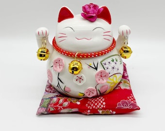 May include: A white ceramic Maneki Neko cat figurine with red accents, a pink flower, and gold bells. The cat sits on a red floral cushion. The cat's collar is red and gold. The cat is a symbol of good luck and fortune.