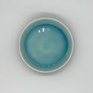 May include: A round ceramic bowl with a light blue interior and a white rim. The bowl's interior has a glossy finish, and the colour transitions from a darker blue at the centre to a lighter shade towards the edges.