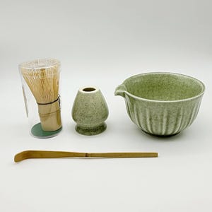 May include: A matcha tea set consisting of a light green ceramic bowl with vertical ridges and a pouring spout, a matching light green ceramic whisk holder, a bamboo whisk inside a clear plastic cylinder, and a long, thin bamboo tea scoop.