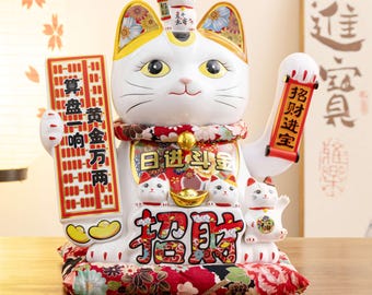 May include: A white ceramic Maneki Neko cat figurine with a red and gold floral collar and base. The cat holds a gold coin and a scroll with Chinese characters. A smaller cat sits on its head.