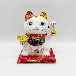 May include: White ceramic Maneki-Neko, or lucky cat, figurine with red and gold accents. The cat is sitting on a red cushion with gold accents and has a gold coin in its left paw and a bell in its right paw. The cat has a red and gold collar with a bell and the word "福" (meaning "good fortune") on its chest.