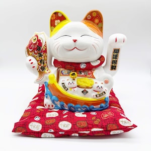 May include: A ceramic Maneki Neko cat figurine with a raised paw, holding a fish. The cat is white with orange and yellow accents, wearing a red collar and sitting on a red patterned cushion. Japanese text is visible.