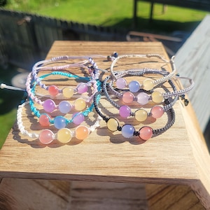 May include: A collection of handmade bracelets with colorful beads. The bracelets feature a variety of cord colors, including white, turquoise, and black. Each bracelet has three round, polished beads in shades of pink, yellow, purple, and blue. The bracelets are displayed on a wooden surface.