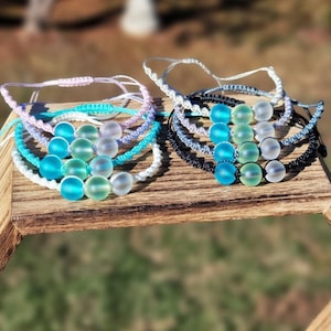 May include: A collection of handmade bracelets featuring frosted glass beads in shades of blue, green, and white. The bracelets are strung on braided cords in various colors, including white, blue, and black. The bracelets are displayed on a wooden surface.
