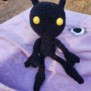 May include: A handmade black crochet doll with yellow eyes. The doll has a rounded head with two small horns and a long, curved antenna. Its arms and legs are outstretched, giving it a unique appearance. The doll is sitting on a purple towel.