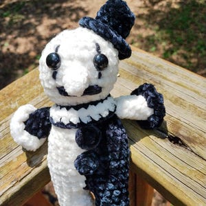 May include: A handmade crochet clown doll in white and navy blue. The doll has a white head with black button eyes, a small nose, and a black mustache. It wears a navy blue top hat and has navy blue and white accents.