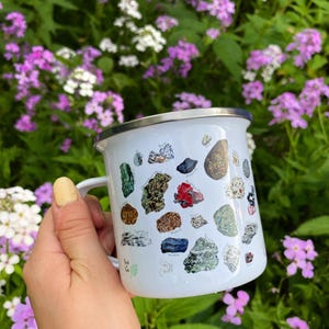 May include: White enamel mug with a colorful illustration of various types of rocks and minerals. The mug has a silver rim and handle.
