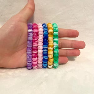 May include: Six beaded bracelets in a variety of colors, including purple, pink, blue, yellow, and green. Each bracelet is made of round beads. The bracelets are stacked together.