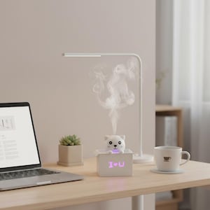 May include: A white cat-shaped essential oil diffuser with the text "I♥U" on a wooden desk. A white desk lamp, a laptop, a small succulent, and a cup of coffee are also on the desk.