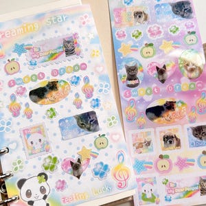 May include: A pink and blue planner with a variety of stickers featuring cats and kittens. The stickers have different designs and phrases such as 'dreaming star', 'sweet dream', 'feeling lucky', 'my dear pencil', and 'cloudy'. The stickers are arranged in a grid pattern on the planner pages.