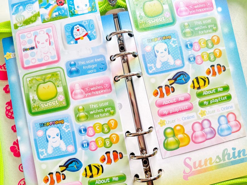 May include: A colorful sticker sheet with various designs, including cartoon characters, apples, and fish. The stickers feature text like "Happy," "Sweet," and "About Me." The sheet is inside a binder.