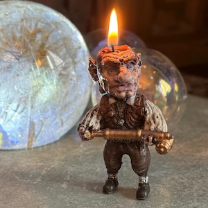 May include: A hand-painted candle sculpture of a goblin holding a small cannon. The goblin is wearing a brown vest and pants and has a lit candle on its head.