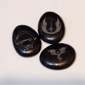 May include: Three black oval-shaped stones with different designs etched into them. The designs are a spaceship, a dragon, and a symbol with wings and a star.