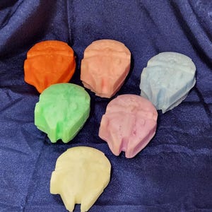 May include: Six colorful soap bars shaped like the Millennium Falcon spaceship from Star Wars. The soaps are in shades of orange, pink, light blue, green, lavender, and yellow.