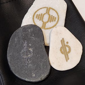 May include: Three smooth, rounded stones with gold symbols on them. The stones are in a black leather pouch.