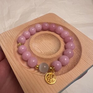 Könnte beinhalten: Ein Armband aus rosafarbenen Edelsteinen mit einem goldenen Anhänger mit chinesischen Schriftzeichen. Das Armband liegt auf einer Holzoberfläche.