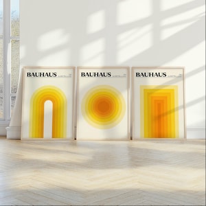 May include: Three framed Bauhaus art prints. Each print features a different geometric design in shades of yellow and orange. The top of each print reads "BAUHAUS" in black text.