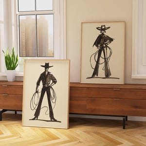 Pu&ograve; includere: Due stampe artistiche incorniciate con illustrazioni in bianco e nero di una persona in un abito ispirato ai cowboy con cappello e corda. Le stampe sono esposte su un mobile in legno.