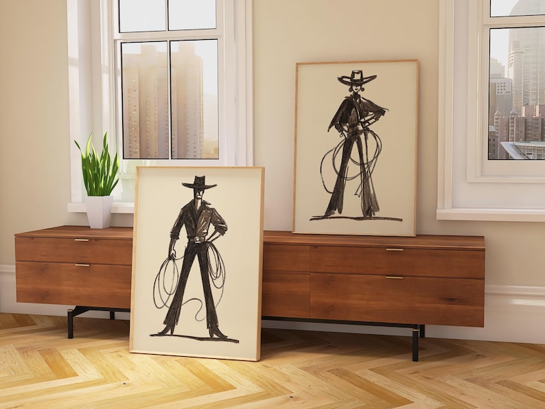 Pu&ograve; includere: Due stampe artistiche incorniciate con illustrazioni in bianco e nero di una persona in un abito ispirato ai cowboy con cappello e corda. Le stampe sono esposte su un mobile in legno.