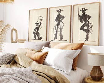 May include: Three framed art prints featuring black ink illustrations of figures in cowboy hats and attire. The artwork hangs above a bed with neutral-toned bedding and decorative pillows. The room has a bohemian aesthetic with a woven lampshade and a bedside table.