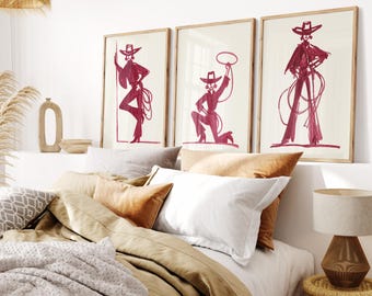 May include: Three framed art prints featuring stylized illustrations of a person in a cowboy hat and attire, rendered in a deep red hue. The prints are displayed above a bed with neutral-toned bedding, including pillows in shades of white, beige, and orange.