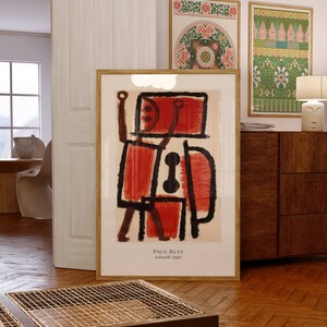 Puede incluir: Una impresión enmarcada de una pintura abstracta roja y negra de Paul Klee, titulada "Adventsbild" (1940). La impresión está colgada en una pared en una habitación con suelo de madera y un armario de madera.
