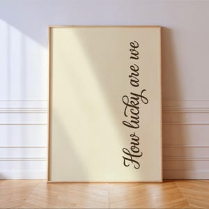 Puede incluir: Impresión enmarcada con la frase "How lucky are we" en escritura cursiva. La impresión tiene un fondo color crema y un marco de madera clara. Está apoyada contra una pared blanca, con un suelo de madera.