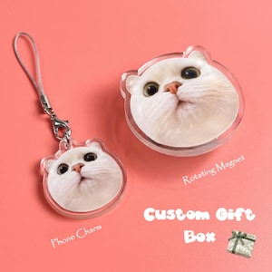 Puede incluir: Un charm de teléfono y un imán giratorio, ambos con la cara de un gato blanco. El charm tiene un cordón plateado. El texto "Custom Gift Box" es visible. Fondo rosa.