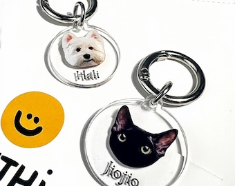 May include: Two circular clear acrylic keychains with silver metal lobster clasps. One keychain features a white dog portrait with the name Hali printed below. The other keychain displays a black cat portrait with the name Jiojio printed below.