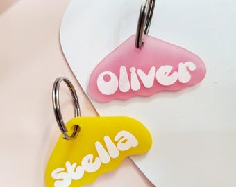 May include: Two colorful plastic pet ID tags in the shape of a cloud with white lettering. The pink tag says "Oliver" and the yellow tag says "Stella".