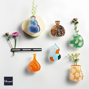 Op de afbeelding: Een verzameling kleurrijke, decoratieve wandvazen in verschillende vormen en patronen. De vazen zijn gevuld met bloemen en groen en zijn bevestigd aan een witte muur. Het woord "Magnet" is linksonder zichtbaar. De tekst "ONCECHO" is ook zichtbaar.