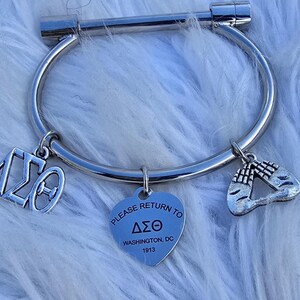 May include: Silver bangle bracelet with three charms. The charms are a heart with the text "PLEASE RETURN TO &Delta;&Sigma;&Theta; WASHINGTON, DC 1913", a Greek letter charm, and a charm with two hands clasped in prayer.