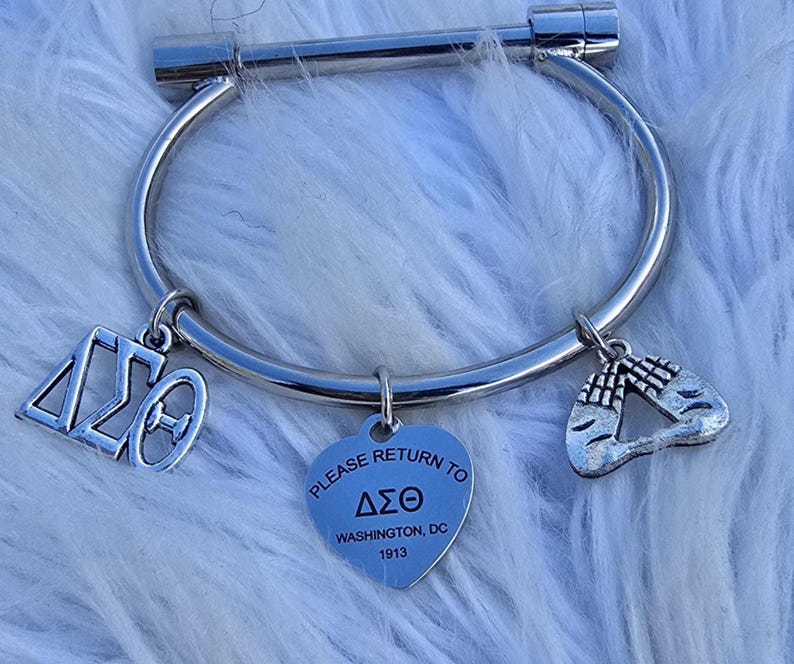 May include: Silver bangle bracelet with three charms. The charms are a heart with the text "PLEASE RETURN TO &Delta;&Sigma;&Theta; WASHINGTON, DC 1913", a Greek letter charm, and a charm with two hands clasped in prayer.