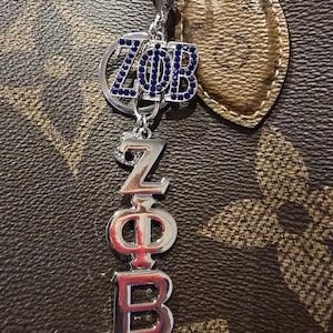 May include: Silver keychain with the letters ZΦB in a combination of blue and silver. The keychain is attached to a brown and tan patterned surface. The letters are embellished with blue rhinestones.