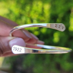 Peut inclure: Un bracelet manchette argenté aux extrémités arrondies. Le bracelet porte l'inscription "ΣΓΡ" gravée à une extrémité et "1922" à l'autre. Le bracelet est présenté sur un fond vert flou.