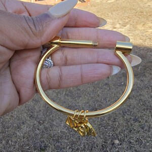 May include: Gold bangle bracelet with a screw-on clasp and a small charm hanging from it. The charm is shaped like a triangle with the letters "19" on it.