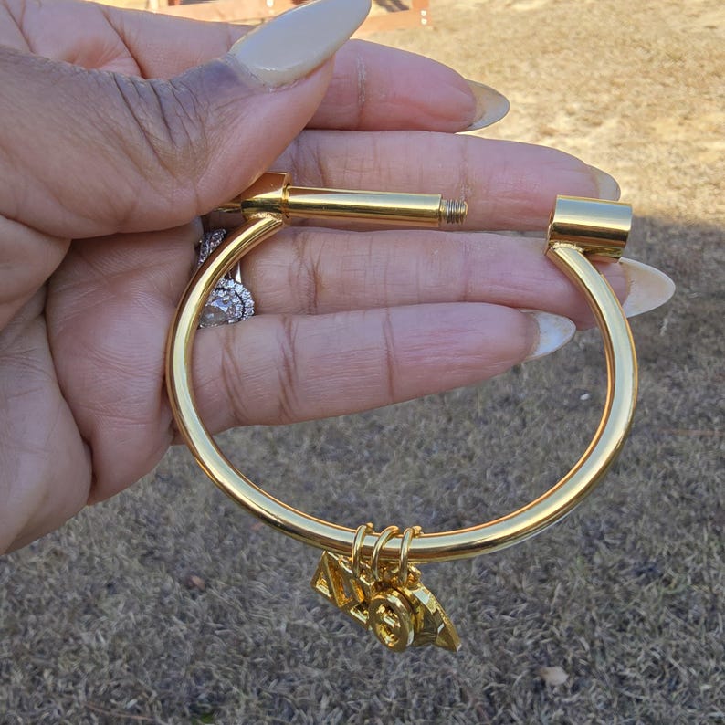 May include: Gold bangle bracelet with a screw-on clasp and a small charm hanging from it. The charm is shaped like a triangle with the letters "19" on it.