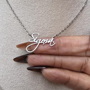 May include: A silver-colored necklace with the name "Sigma" in a cursive font. The pendant hangs from a delicate chain. The necklace is displayed against a neutral background.