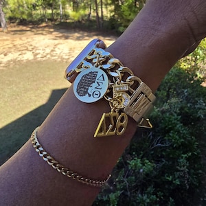 May include: A gold chain bracelet with a charm featuring a black silhouette of a woman's head with the text "I AM" and the Greek letters "ΔΣΘ". The bracelet also has a charm with the number "5" and rhinestones.