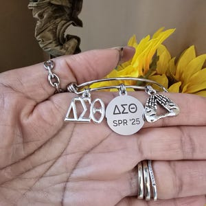 May include: A silver charm bracelet with three charms. The charms are a sorority symbol, a round charm with the sorority symbol and the text "SPR '25", and a charm with two hands clasped together in prayer.